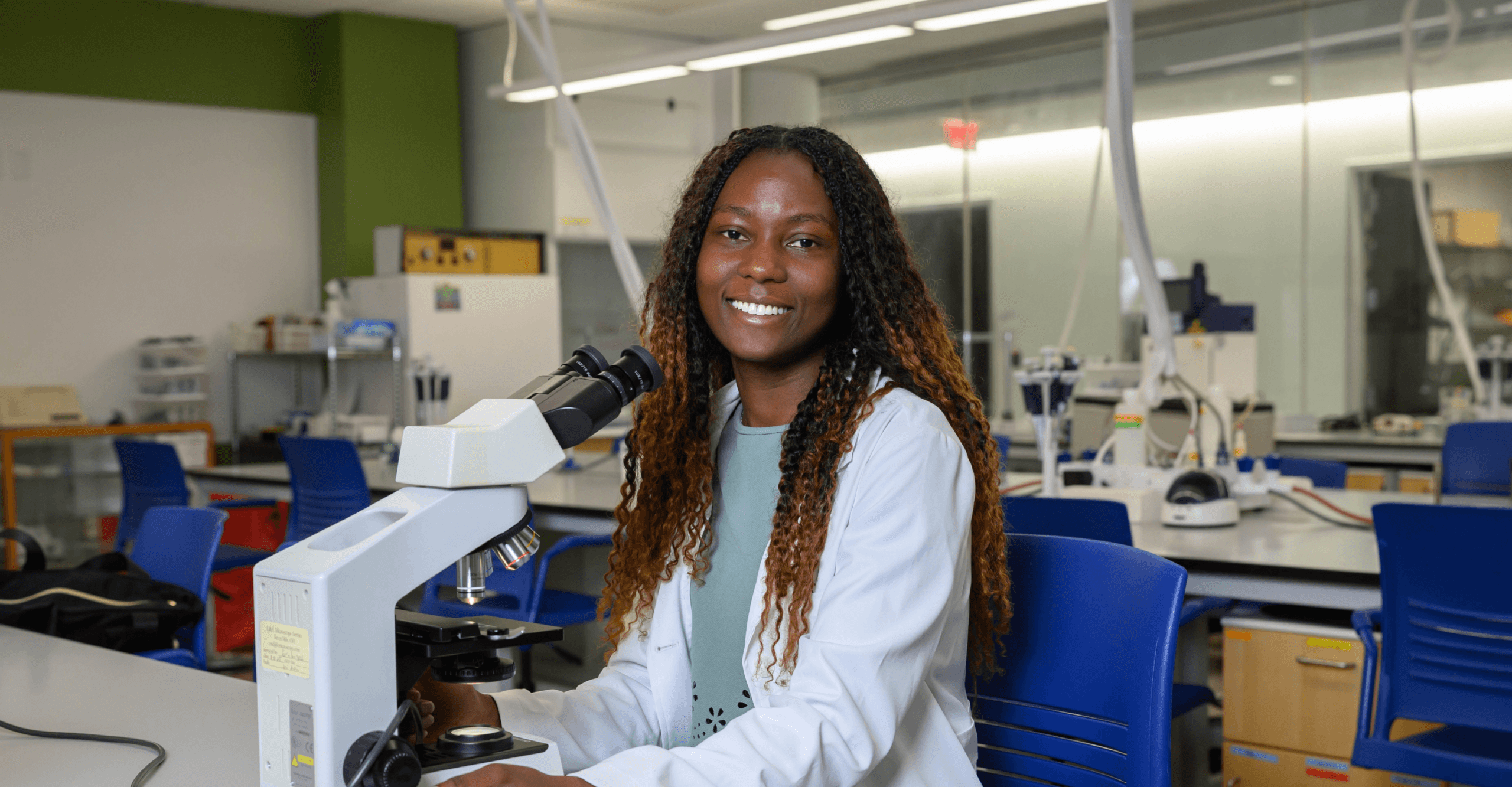 Smiling woman wearing a lab coat sitting at a table with a microscope